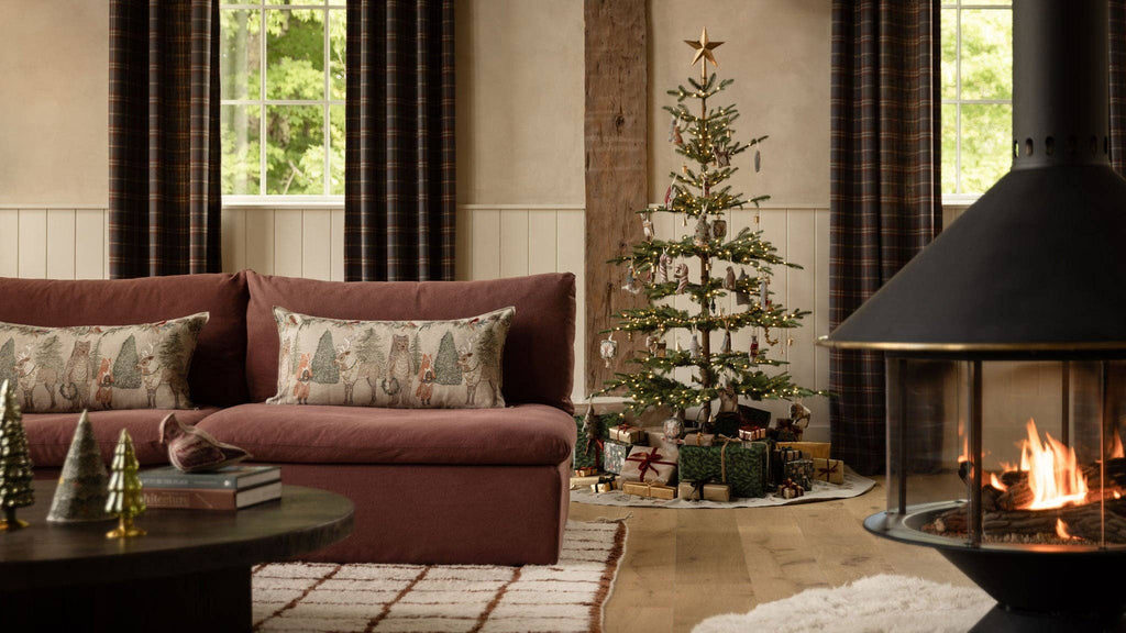 Coral & Tusk Christmas Cheer Lumbar Pillow, a rectangular embroidered holiday pillow featuring woodland animals and Christmas trees on a sofa next to a decorated tree and fireplace.