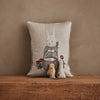 The Coral & Tusk Easter Bunny Pocket Pillow, featuring an embroidered rabbit in charming clothes among flowers, rests on a wooden surface with three soft toys—a rabbit, chick, and bunny—creating a delightful Easter display.