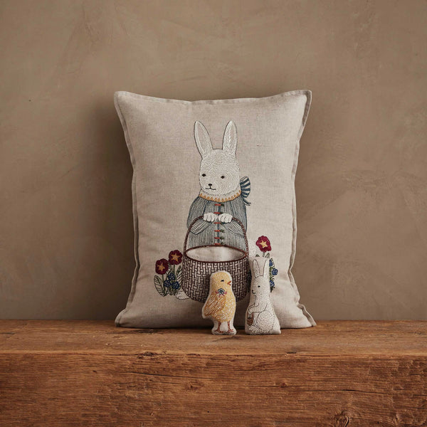 The Coral & Tusk Easter Bunny Pocket Pillow, featuring an embroidered rabbit in charming clothes among flowers, rests on a wooden surface with three soft toys—a rabbit, chick, and bunny—creating a delightful Easter display.