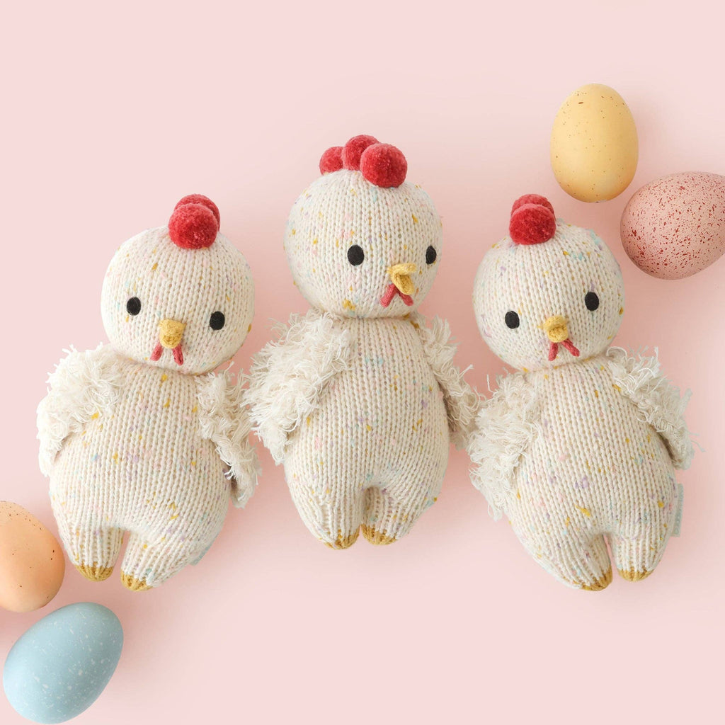 Three Cuddle + Kind Baby Chicken plushies—heirloom-quality and hand-knit in Peru with red combs and yellow beaks—are arranged side by side on a pink background among pastel speckled eggs.