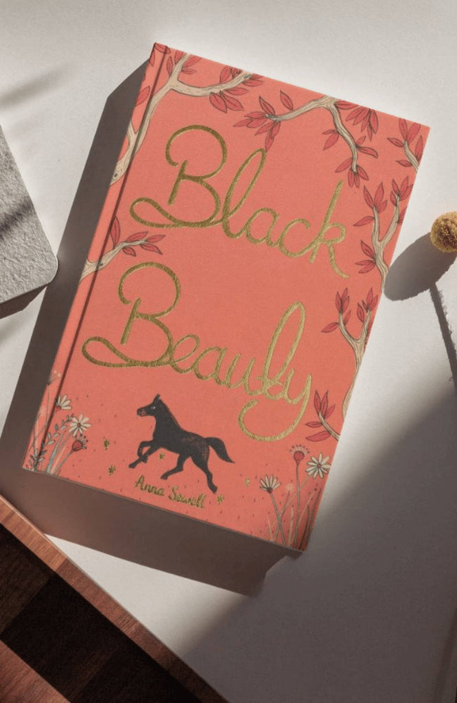 The "Black Beauty | Collector's Edition | Hardcover" features a pink and gold cover with a black horse silhouette and floral accents, resting on a white surface as sunlight casts gentle shadows across its elegant design.