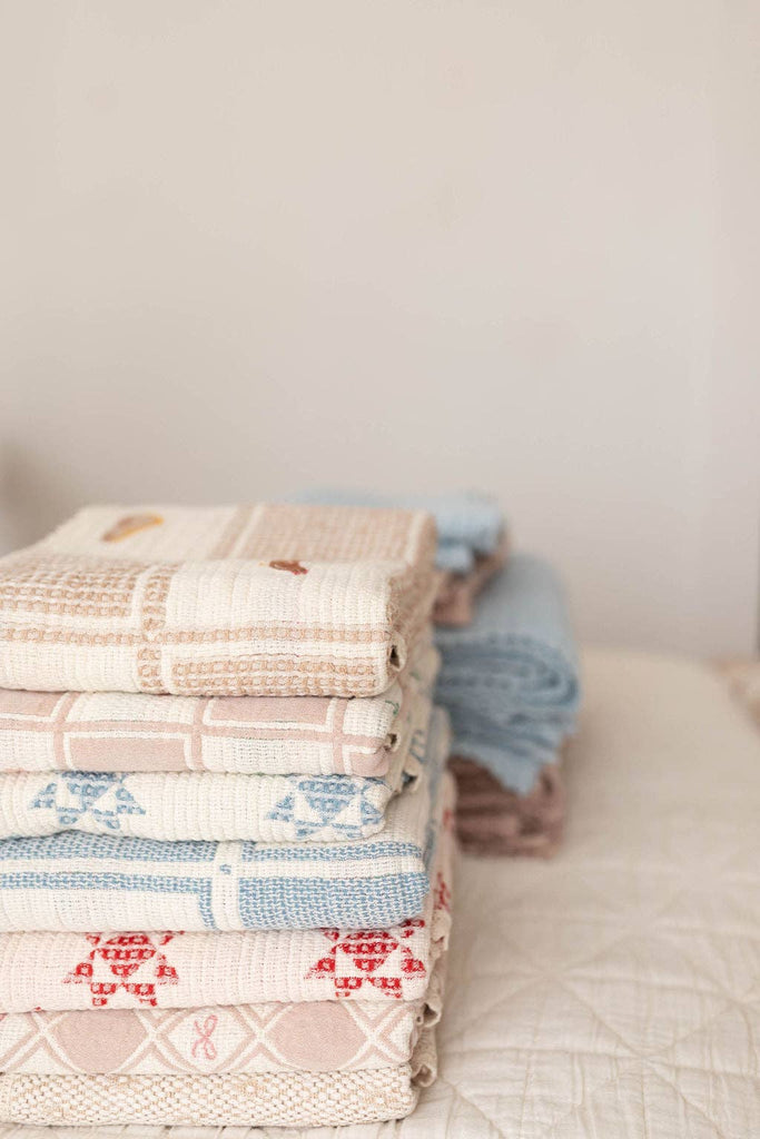 The Organic Cotton Baby Blanket - Farm, featuring soft pastel patterns and delicate crochet trim, is neatly folded on a bed with a white quilted cover against a light, neutral background.