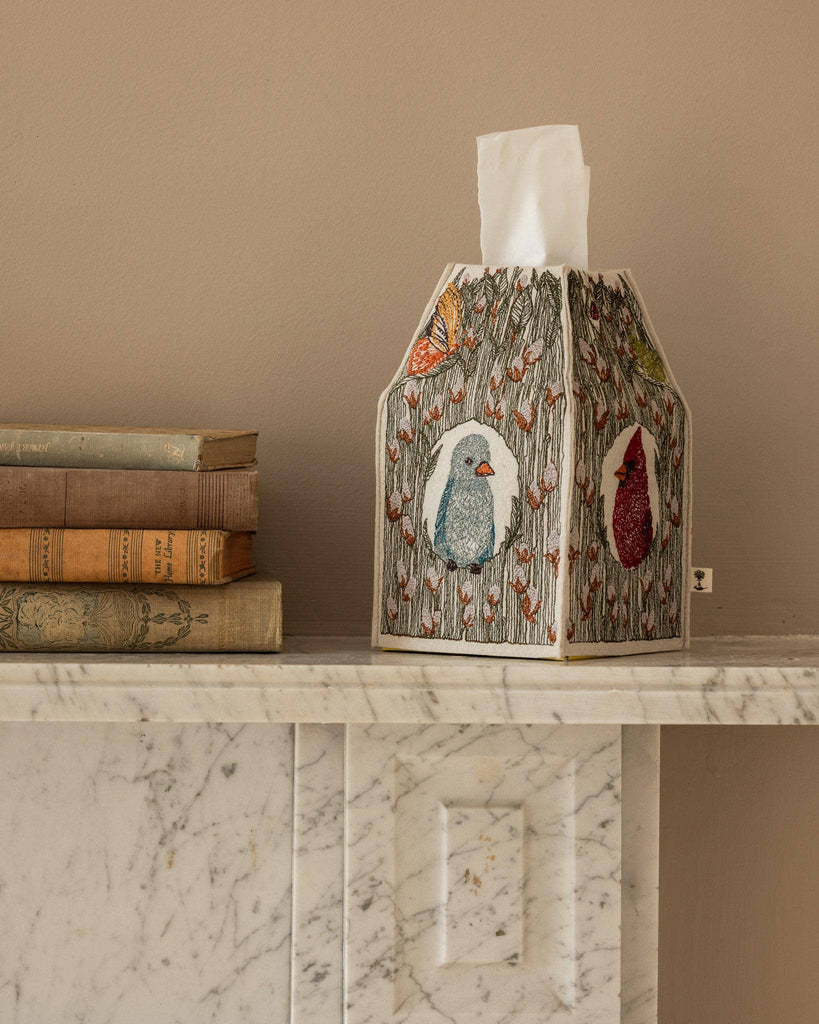 The Coral & Tusk Birdhouse Tissue Box Cover with embroidered birds and flowers sits on a white marble mantel beside vintage books against a beige wall.