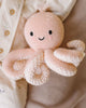 The Cuddle + Kind Baby Octopus (Soft Coral Pink) is a soft, hand-knit toy made from heirloom-quality Peruvian cotton yarn, featuring a smiling face and black eyes, resting against cream-colored fabric with buttons.