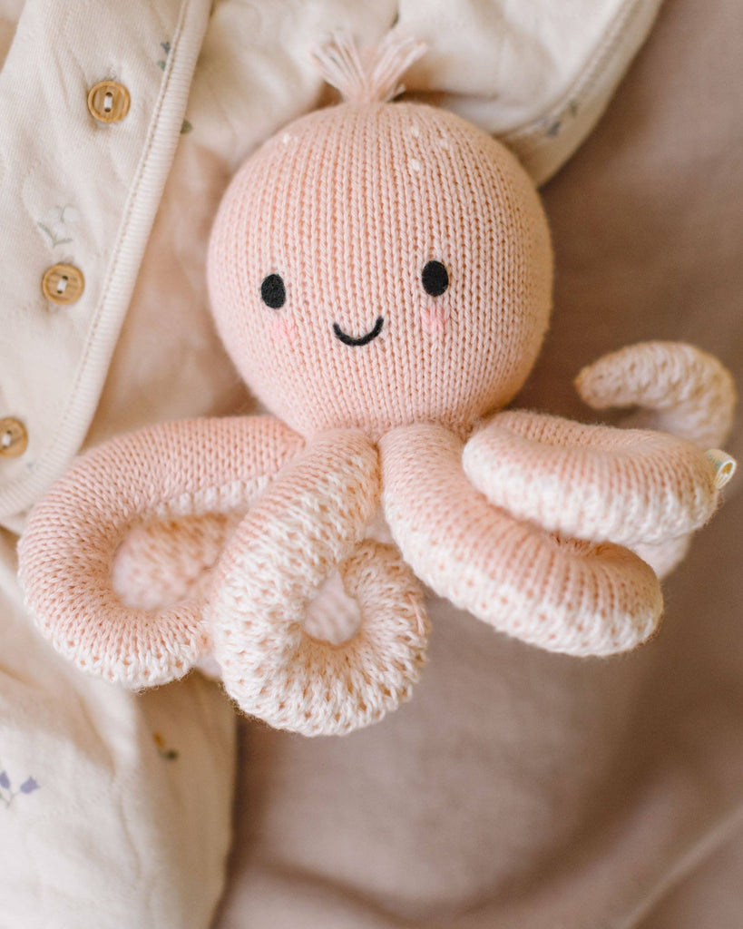 The Cuddle + Kind Baby Octopus (Soft Coral Pink) is a soft, hand-knit toy made from heirloom-quality Peruvian cotton yarn, featuring a smiling face and black eyes, resting against cream-colored fabric with buttons.