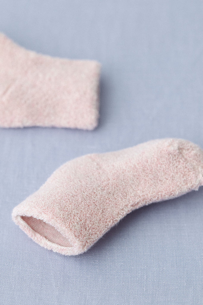 A close-up of a soft, fuzzy Baby Pile Knit Sock - Pink lying on a light blue fabric surface. Another matching pink sock is partially visible in the upper left corner of the image. The fluffy socks appear warm and cozy, suitable for keeping feet warm and comfortable.