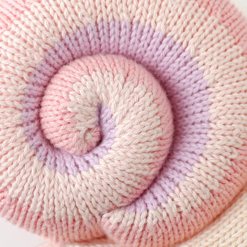 Close-up of the Cuddle + Kind Baby Snail (Peony), showing hand-knit fabric rolled in a spiral with pink, lavender, and cream stripes. Its cozy texture recalls the softness of our collectible, handmade-in-Peru dolls.