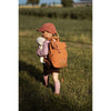 A young child outdoors on a sunny day, wearing a pink cap and socks with a brown backpack, walks on grass holding the Senger Naturwelt Stuffed Animal - Small Sheep. The child is facing away from the camera.