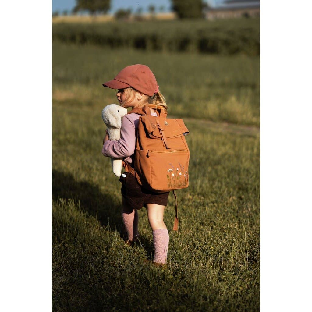 A young child outdoors on a sunny day, wearing a pink cap and socks with a brown backpack, walks on grass holding the Senger Naturwelt Stuffed Animal - Small Sheep. The child is facing away from the camera.
