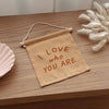 The I Love Who You Are Canvas Sign - Peach, a small handmade natural canvas sign with peach lettering, rests on a wooden surface beside a decorative shell dish and white dried flowers.