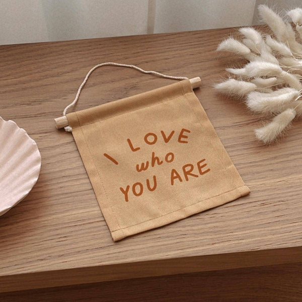 The I Love Who You Are Canvas Sign - Peach, a small handmade natural canvas sign with peach lettering, rests on a wooden surface beside a decorative shell dish and white dried flowers.