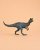 The Schleich Dilophosaurus, a small blue and green dinosaur figurine with a ridged back and short arms from the DINOSAURS collection, stands on a plain peach background, capturing the charm of prehistoric giants.