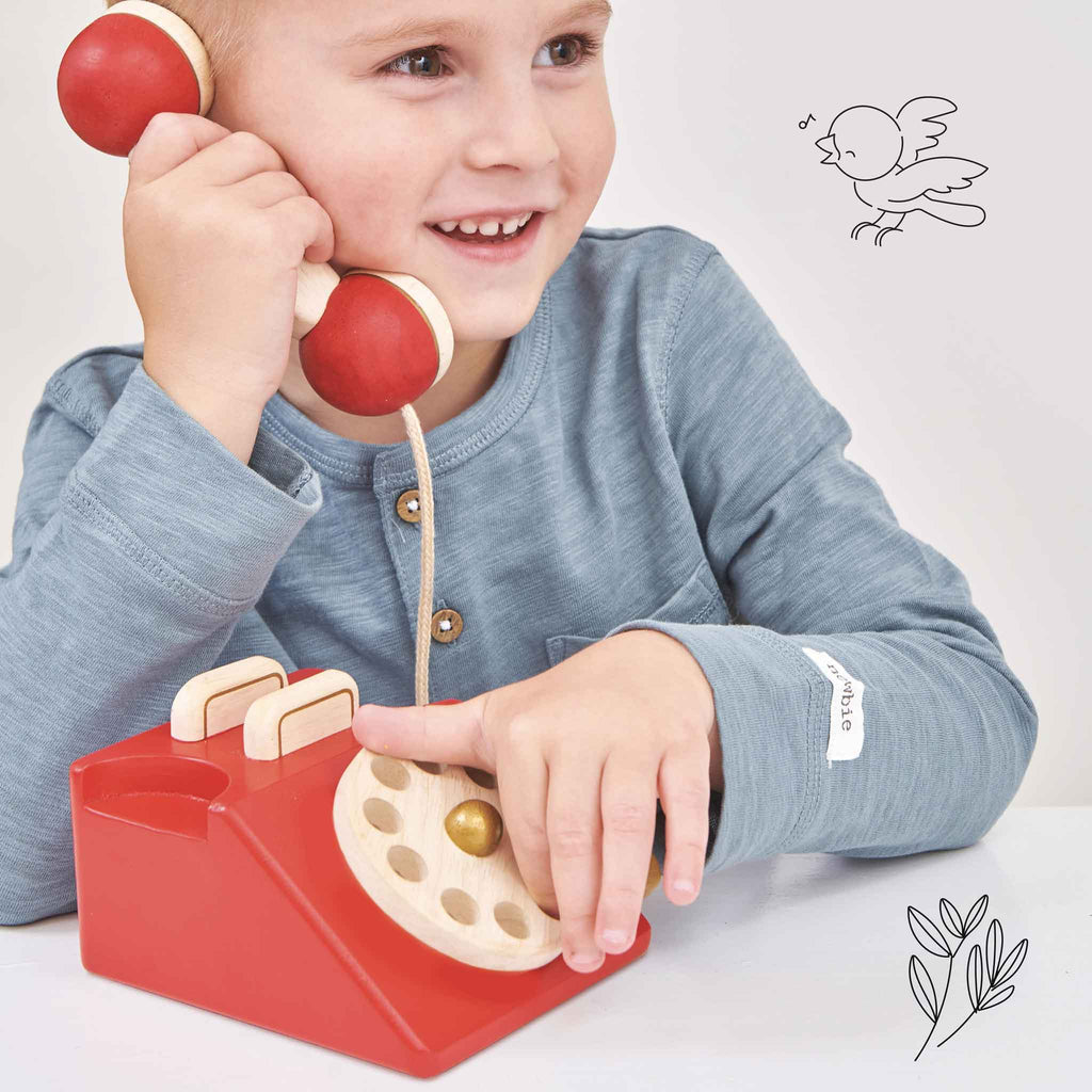 A smiling child in a blue shirt holds the Vintage Wooden Telephone to their ear, with simple line drawings of a bird and leaves in the background.