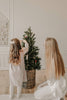 A young girl and a woman decorate a small Christmas tree in a wicker basket with Sabo Concept Christmas Ornaments. The girl hangs the decorations while the woman kneels beside her in a bright room with wooden floors.