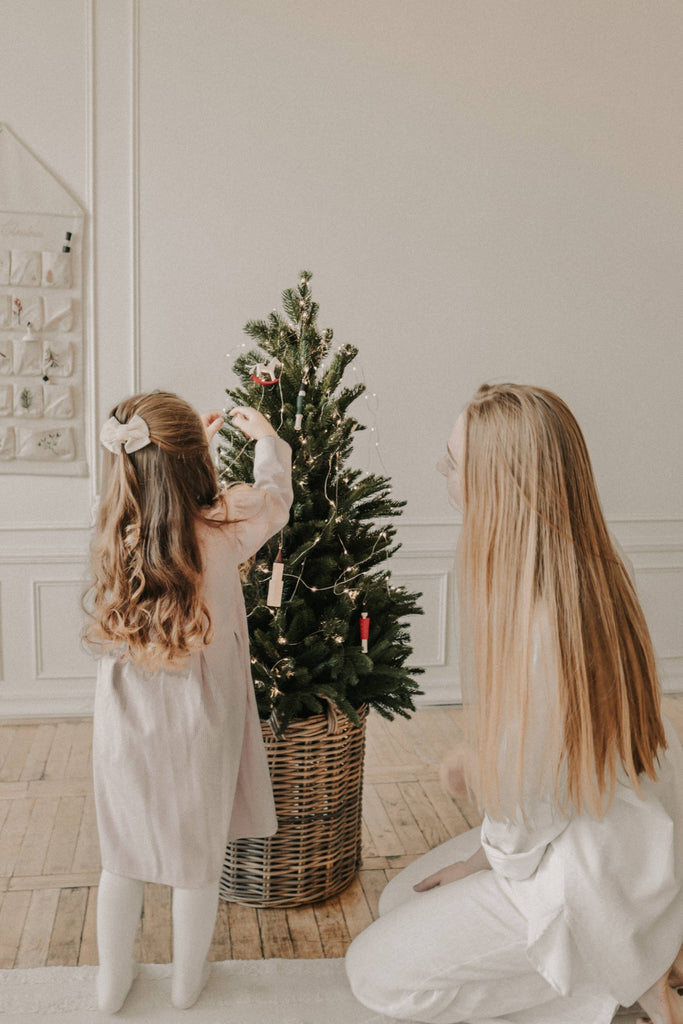 A young girl and a woman decorate a small Christmas tree in a wicker basket with Sabo Concept Christmas Ornaments. The girl hangs the decorations while the woman kneels beside her in a bright room with wooden floors.