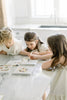 Three young girls sit at a marble kitchen counter, painting with watercolors and their Wishy Washy (Water Reveal Edition) book in natural daylight. Art supplies and the reusable water reveal book are open in front of them.