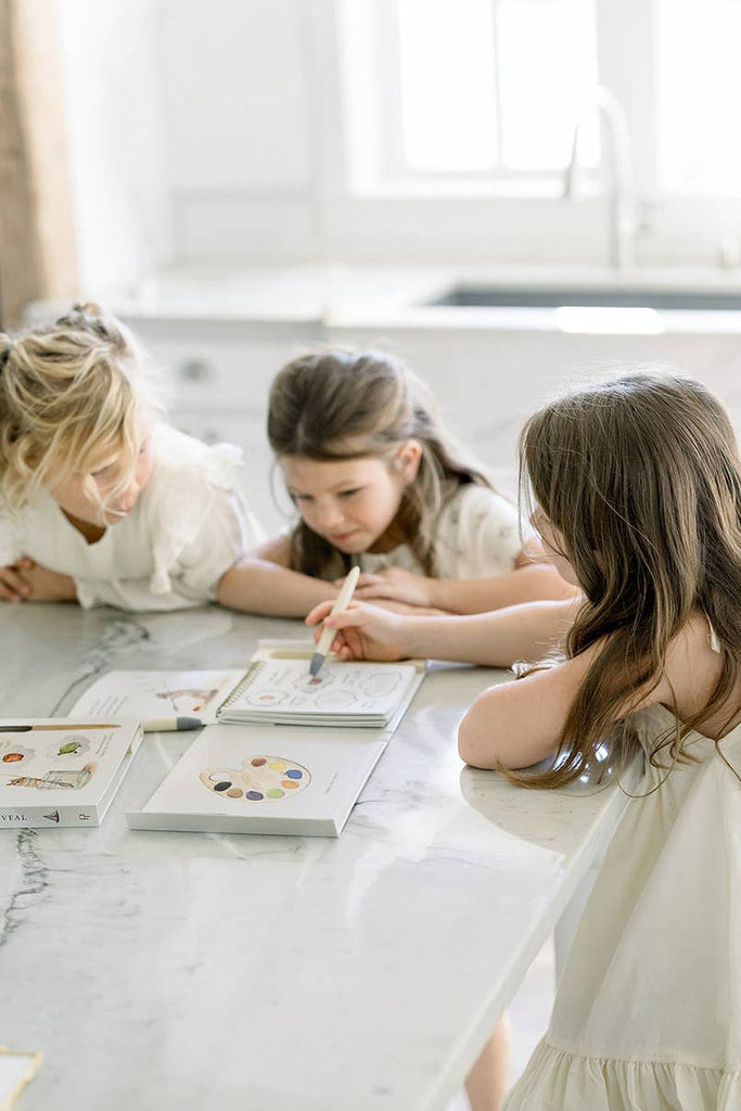 Three young girls sit at a marble kitchen counter, painting with watercolors and their Wishy Washy (Water Reveal Edition) book in natural daylight. Art supplies and the reusable water reveal book are open in front of them.