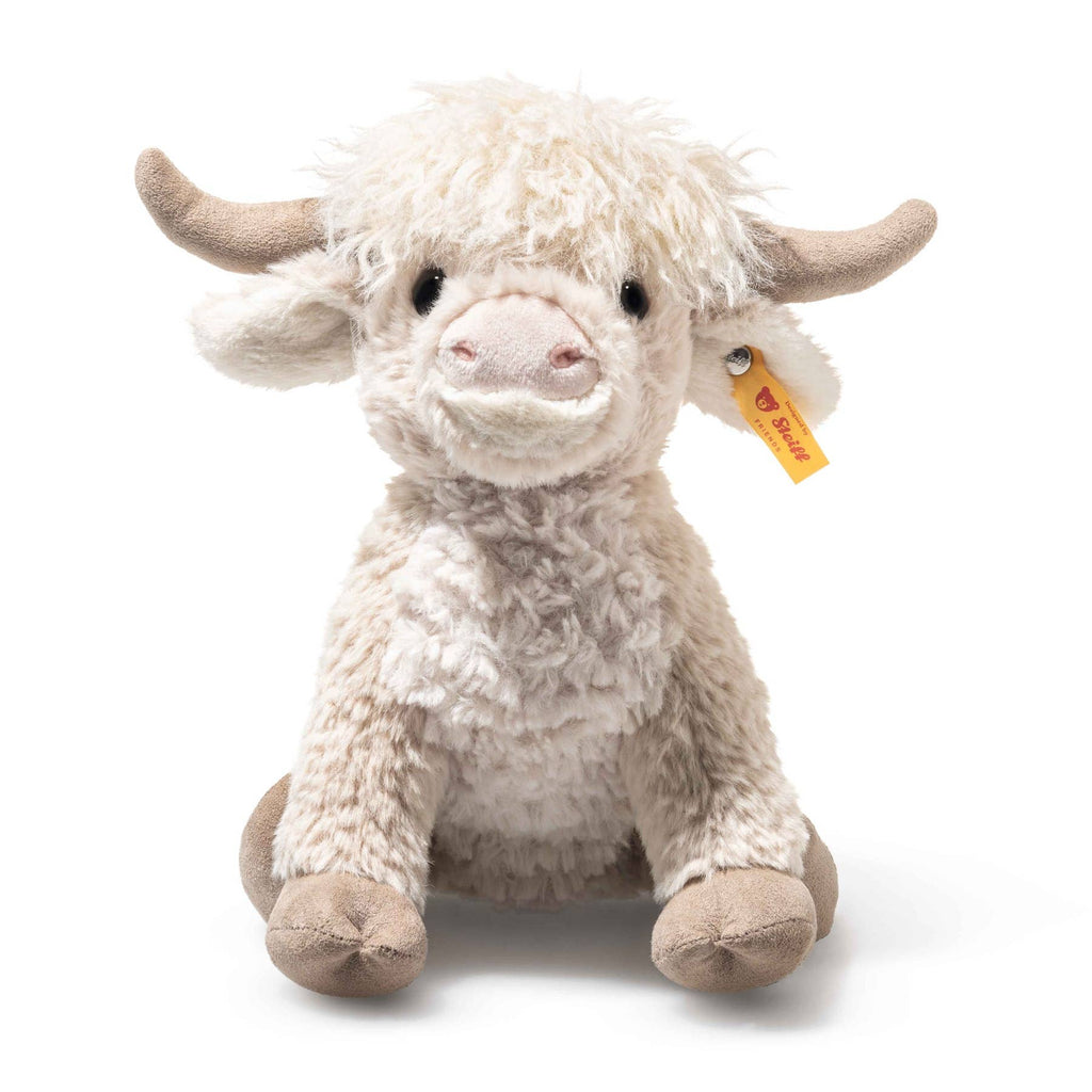The Steiff Harper Highland Cow is a plush toy with light brown and cream shaggy fur, curved horns, and a yellow ear tag, sitting upright—perfect as a nursery companion.