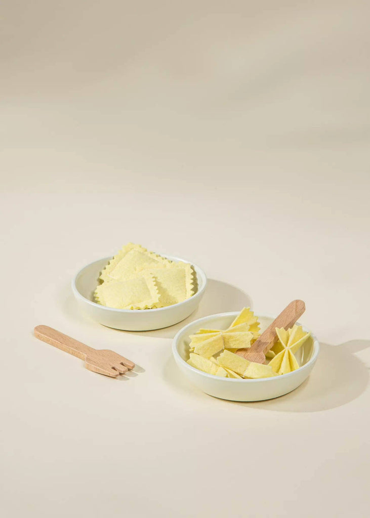 On a light beige surface, two small bowls showcase the delightful offerings from the Wooden Pasta Playset: one brimming with square ravioli and the other with triangular pasta. Accompanying them are wooden utensils, ideal for imaginative play sessions.