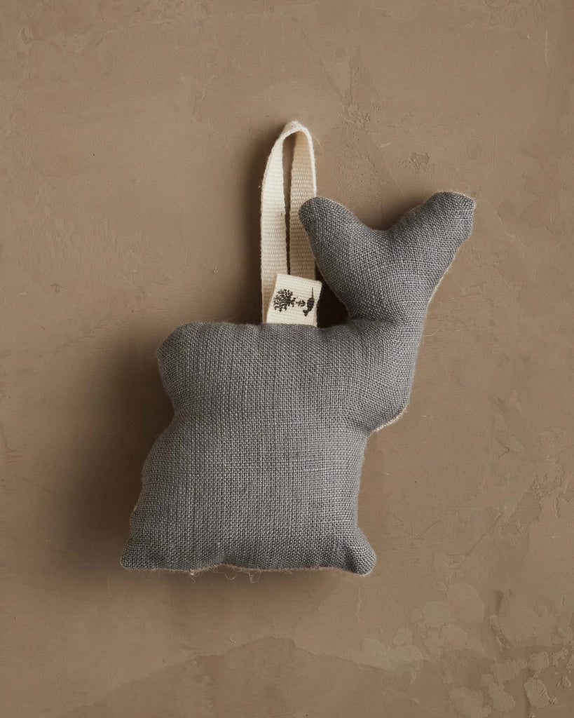 The Coral & Tusk Deer with Lights Ornament is a small, gray fabric deer with a white hanging loop, perfect for adding woodland charm to your Christmas tree, shown against a textured beige wall.