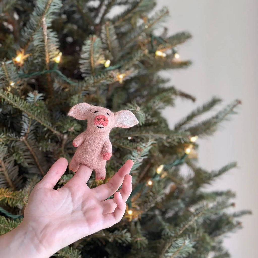 A hand showcases a handmade pink "Pig Finger Puppet" with big ears, ideal for storytelling, against the backdrop of a Christmas tree sparkling with lights.