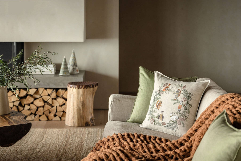 A cozy living room with a beige sofa, green nature-themed pillows, the Coral & Tusk Tree Trimmers Wreath Pillow, a chunky brown knit blanket, a small wooden stump table, stacked firewood, and decorative tree ornaments on a shelf.
