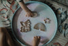 Two children’s hands play with wooden shapes of a unicorn, rainbow, star, and cloud—using the Unicorn Cutter Set: Eco Playdough Tool—on a pastel circular tray atop a floral-patterned blanket, creating the perfect sensory play scene.