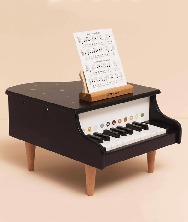 The Wooden Grand Piano Toy is a small black piano with wooden legs and color-coded keys, made from FSC®-certified wood. It includes a music sheet on a stand above the keys, shown against a plain light beige background.