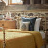 A rustic bedroom with a wooden bed, mustard quilt, plaid pillows, the Coral & Tusk Bat Wing Surprise Pillow, a bat plush, stone wall, and a nightstand with a ghost basket and pumpkin creates a cozy Halloween vibe.