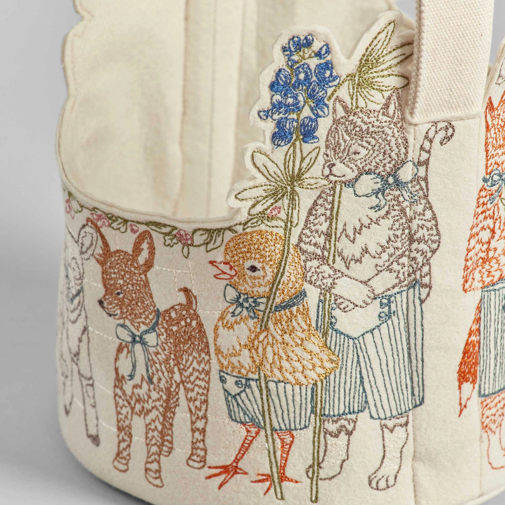 The Coral & Tusk Easter Parade Basket showcases embroidered woodland creatures like a cat and bird, dressed in outfits and holding flowers. Detailed stitching enhances its felt interior, offering a charming handmade look against a plain light gray surface.