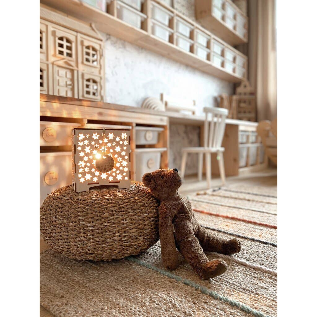 A Senger Naturwelt Stuffed Animal - Large Brown Bear leans on a woven ottoman atop a striped rug, next to a glowing lamp with star cutouts. Wooden shelves, toys, and sunlight from the window make the room warm and inviting.