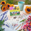 The Magical Minds Garden Kit is displayed on a white table with sunflowers, snapdragons, and lavender. This fun STEM DIY kit includes plant markers, seed sticks, wooden butterfly decorations, and a blue cup—ideal for creating a pollinator habitat.
