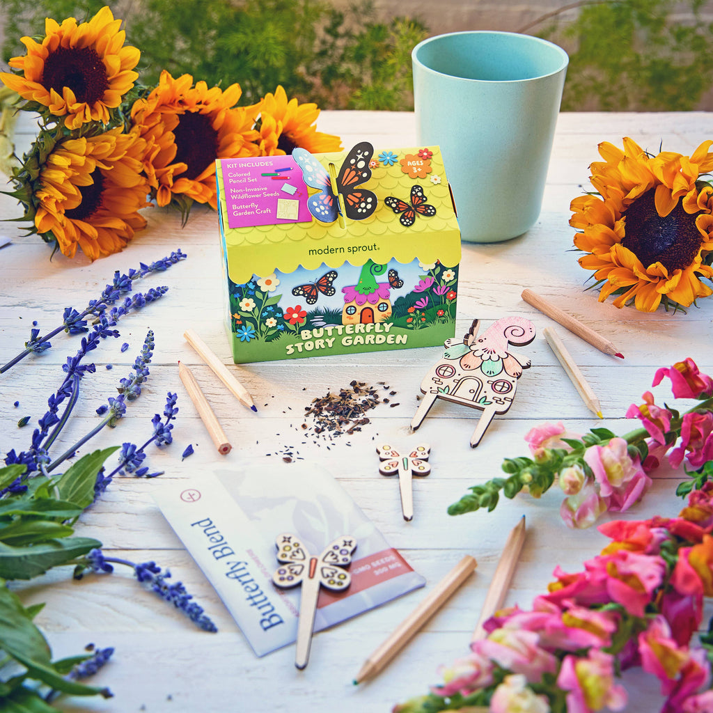 The Magical Minds Garden Kit is displayed on a white table with sunflowers, snapdragons, and lavender. This fun STEM DIY kit includes plant markers, seed sticks, wooden butterfly decorations, and a blue cup—ideal for creating a pollinator habitat.