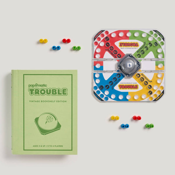 The Board Game: Trouble Vintage Bookshelf Edition features a green book-shaped box, colorful Pop-O-Matic die roller, and red, blue, yellow, and green playing pegs displayed on a light background.