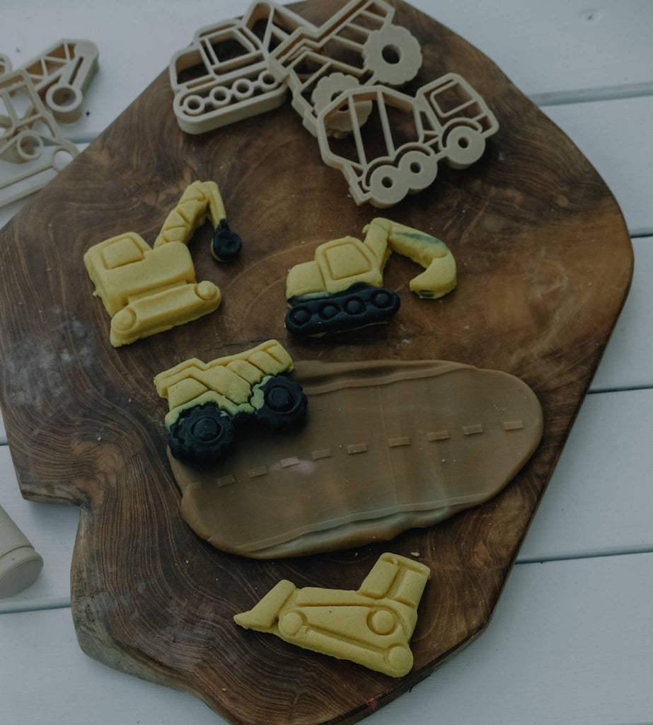 Cookies shaped like construction vehicles and their matching plastic cutters from the Construction Cutter Set – Eco Playdough Tool are arranged on a wooden board, featuring a road-shaped cookie in the center. Ideal for sensory play, these cookies and eco-friendly tools are displayed against a clean white wooden background.