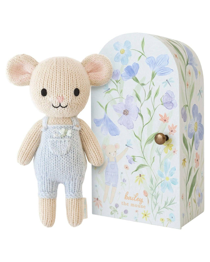 Cuddle + Kind Tiny Bailey the Mouse, fair trade and handmade in Peru, comes in light blue overalls and stands beside a decorative box with pastel flowers, an illustration of Bailey, and “bailey the mouse” text.