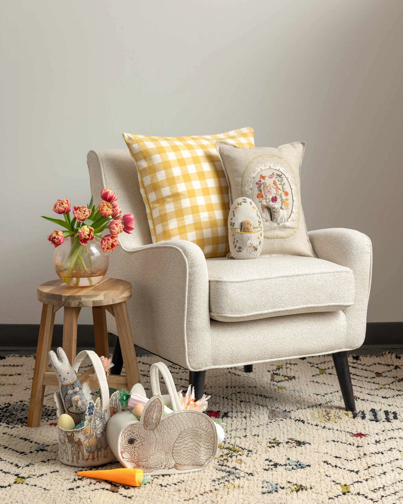 A cozy armchair features the Coral & Tusk Sugar Egg Pocket Pillow in yellow plaid and florals, beside a wooden stool with a vase of tulips. Bunny baskets with eggs and toys sit on a patterned rug, creating festive Easter décor.