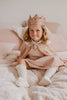 A young girl wearing the Cape - Pink sequins, a sparkly pink crown, a dress, and knee-high socks smiles while sitting on a bed with light pink and white bedding.