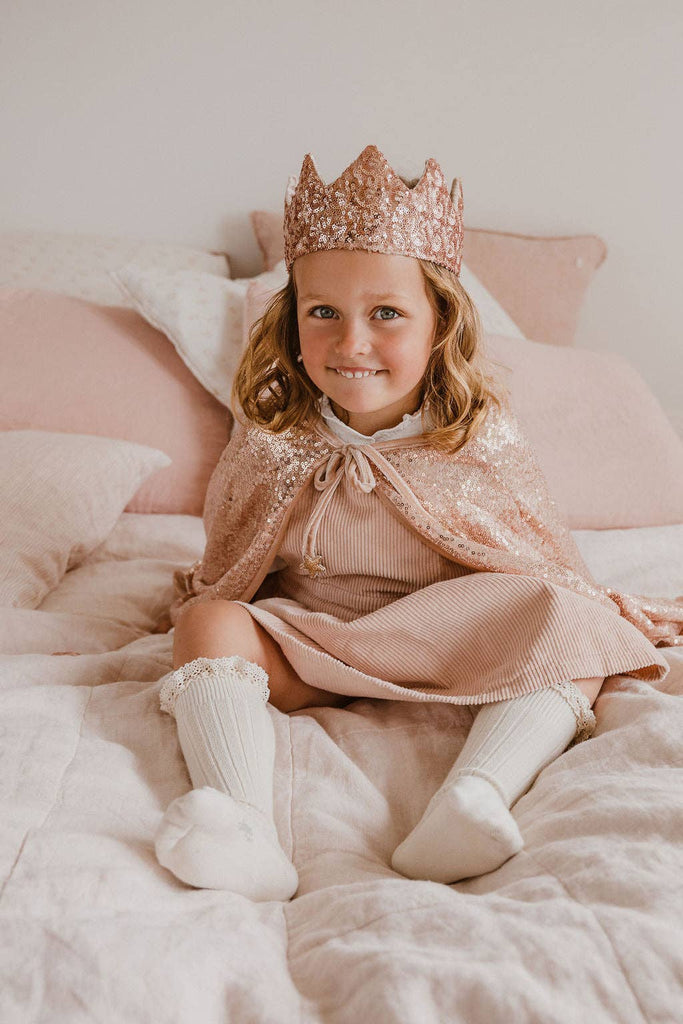 A young girl wearing the Cape - Pink sequins, a sparkly pink crown, a dress, and knee-high socks smiles while sitting on a bed with light pink and white bedding.