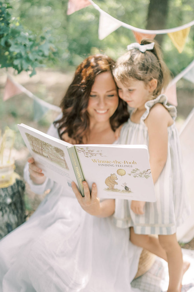 A woman and a young girl smile while reading Winnie-the-Pooh: Finding Feelings board book together outdoors, surrounded by trees and colorful bunting, exploring emotions for toddlers.