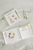 The Wishy Washy (Water Reveal Edition) reusable activity book is open on a marble surface, showing a paint palette illustration that says “SWISH SWISH.” This toddler-friendly travel set includes two water brushes and a book with colorful art supplies.