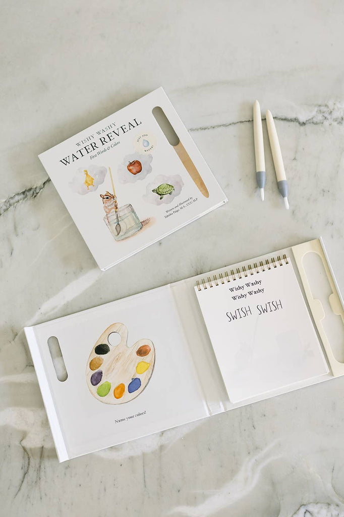 The Wishy Washy (Water Reveal Edition) reusable activity book is open on a marble surface, showing a paint palette illustration that says “SWISH SWISH.” This toddler-friendly travel set includes two water brushes and a book with colorful art supplies.
