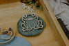 A wooden frog-shaped cookie cutter from the Pond Life Cutter Set – Eco Playdough Tool rests on green playdough, framed by the edges of a wooden tray, with artificial flowers and a striped cloth nearby.