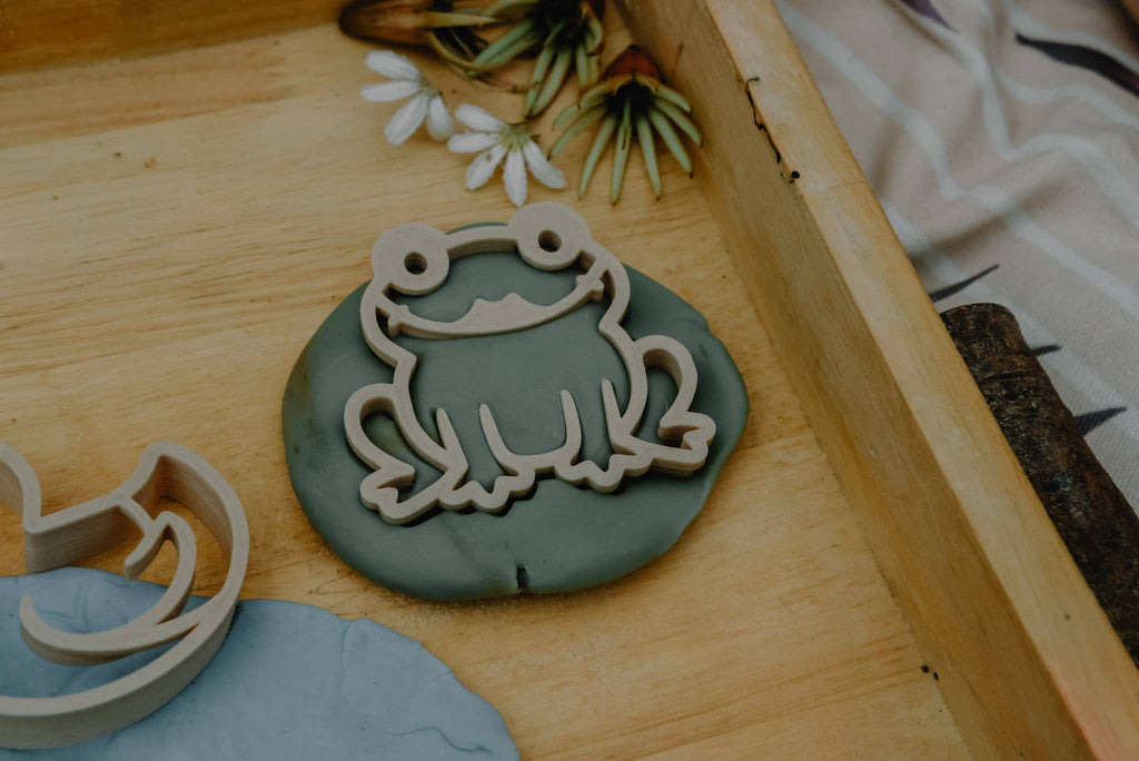 A wooden frog-shaped cookie cutter from the Pond Life Cutter Set – Eco Playdough Tool rests on green playdough, framed by the edges of a wooden tray, with artificial flowers and a striped cloth nearby.