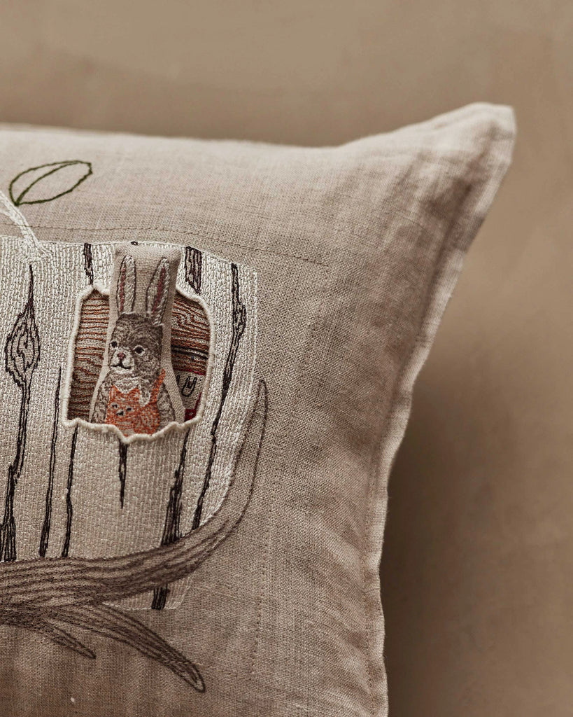 A close-up of the Coral & Tusk Aspen Log Bunnies Pocket Pillow shows a beige linen pillow embroidered with tree trunks and a removable rabbit in an orange patterned shirt peeking from a tree hollow.