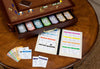 The Monopoly Luxury Edition board game sits on a wooden table, with organized trays for money and cards, die-cast metal pieces, and an open rulebook showing game info and property details.