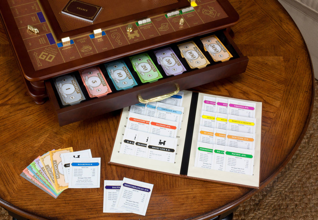 The Monopoly Luxury Edition board game sits on a wooden table, with organized trays for money and cards, die-cast metal pieces, and an open rulebook showing game info and property details.