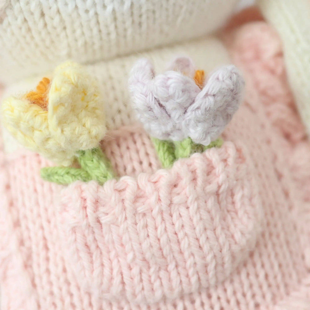 Cuddle + Kind Clover the Bunny (Blossom) features two crocheted flowers—one yellow, one pale purple—nestled in a pink knitted pocket against white and pink handmade Peruvian fabric, showcasing beautiful craftsmanship.