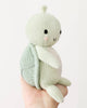 A hand holds the Cuddle + Kind Baby Sea Turtle (Kelp Green), an heirloom-quality, hand-knit plush with a light green body, round shell, black eyes, a small smile, and pink cheeks against a plain white background.