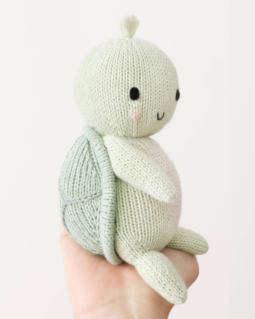 A hand holds the Cuddle + Kind Baby Sea Turtle (Kelp Green), an heirloom-quality, hand-knit plush with a light green body, round shell, black eyes, a small smile, and pink cheeks against a plain white background.